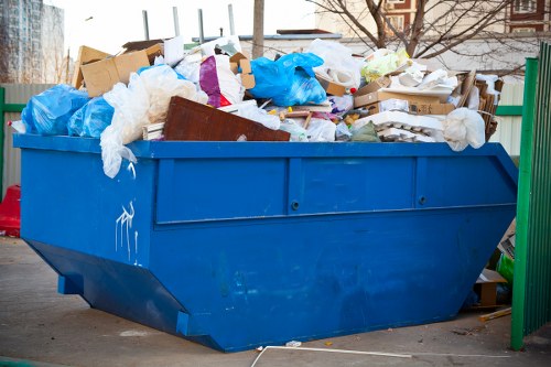 A large blue skip bin filled with mixed waste materials, including cardboard boxes, white and blue plastic bags, crumpled paper, and pieces of white foam, is situated outdoors on a paved area. The bin is positioned near a green metal fence on the right side and appears to be in a residential or commercial setting, with some trees and buildings visible in the background. The waste items are piled high above the top edge of the bin, indicating it is full or nearly full. The skip's surface shows some scuff marks and scratches, and the environment looks clear and well-lit, suggesting daytime. This image visually represents rubbish collection and waste removal services, akin to those provided by Commercial Waste Stockwell serving the local London area, including nearby postcode regions.