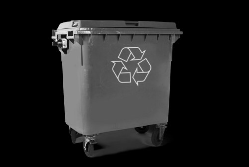A large, dark grey plastic wheelie bin with a hinged lid, prominently displaying a white recycling symbol on its front. The bin is positioned on a concrete or paved surface, with small caster wheels visible at the base, indicating mobility. The background is plain and dark, drawing focus to the bin itself. This bin appears suitable for general waste collection or recycling purposes and is representative of the waste management services offered by Commercial Waste Stockwell in the London area, close to Stockwell. The photograph emphasizes the bin's sturdy, textured plastic surface and practical design, suitable for exterior use on a driveway or street during rubbish removal operations.
