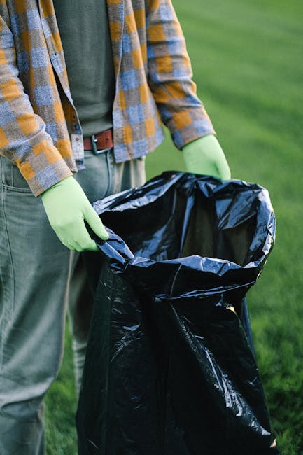 The image shows a person standing outdoors on a grassy area, holding a large black rubbish bag open with both hands. The person is wearing a plaid shirt with blue, yellow, and gray tones, light-colore