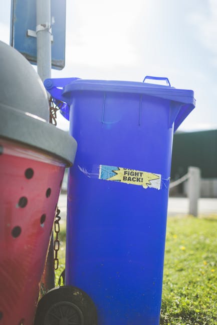 A tall, bright blue wheeled rubbish bin with a partially open lid is positioned on a grassy area outdoors. The bin has a small yellow sticker with blue text that reads 'FIGHT BACK!', and it appears to