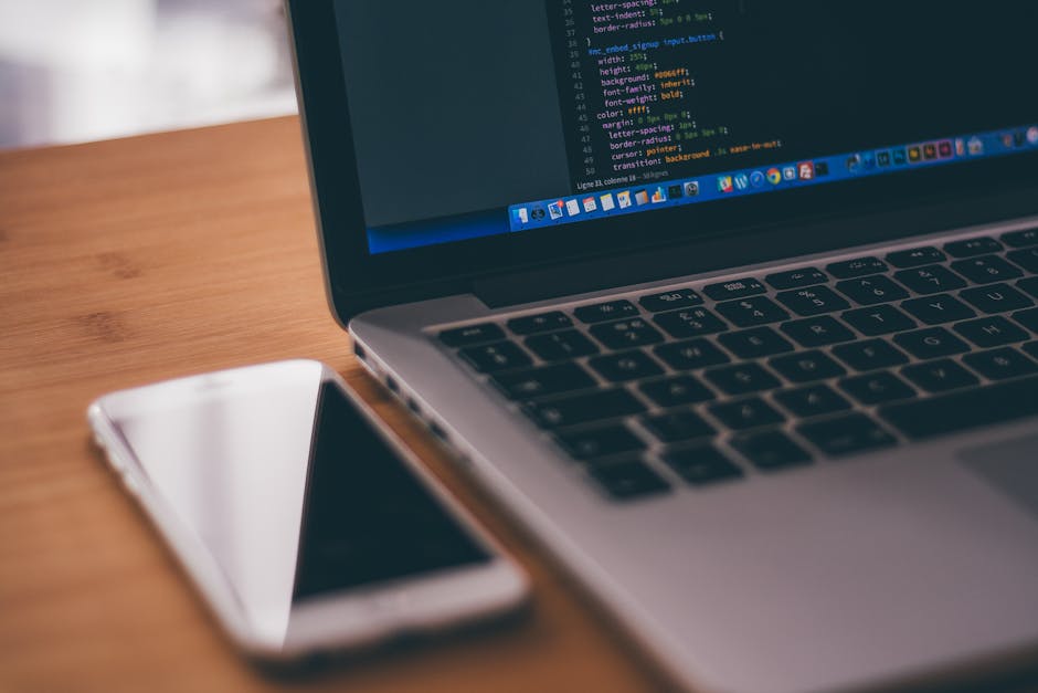 The image depicts a close-up view of a laptop placed on a wooden desk, with its screen displaying lines of code in a dark-themed programming interface. Next to the laptop, on the right side, there is 