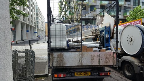The image shows a flatbed truck parked at the side of an urban street, loaded with various types of rubbish including white and beige panels, metal frames, and miscellaneous debris, some leaning against the truck's side. The truck appears to be used for waste collection, with a metal cage structure partially enclosing the load to secure the rubbish. The vehicle's license plate, 'NX12 GHA,' is visible at the rear. In the background, modern multi-storey residential and commercial buildings can be seen, along with a traffic light at an intersection. The scene is set during daylight hours with overcast weather. The environment suggests a typical urban setting that might require rubbish removal services from companies like Commercial Waste Stockwell to manage waste collection in the area.
