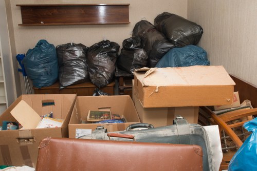 The image shows an indoor storage space or a room used for rubbish collection, featuring several black and blue plastic rubbish bags piled against a wall, with some bags appearing full and tightly knotted. In the foreground, there are open cardboard boxes filled with miscellaneous waste, including paper and small packaging items. A large cardboard box with a slightly torn corner rests atop the boxes, indicating it is partially damaged. To the right, a metallic object, possibly part of a cleaning or garden tool, lies next to a wooden or metal frame, which could be part of a chair or small shelving unit. The background reveals a wooden shelf mounted on the wall, with a plain beige or light brown wall beneath it. The overall scene appears cluttered, with waste items casually stacked, suggesting a temporary collection area before removal. This setting aligns with the services provided by Commercial Waste Stockwell in managing rubbish within the Stockwell area, south London, highlighting the type of waste typically handled by commercial rubbish removal services.